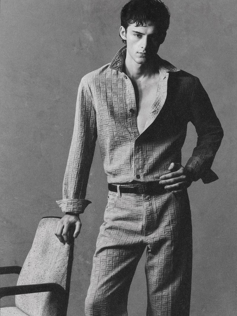 Monochrome portrait of a male model in an unbuttoned patterned shirt and matching pants standing next to a chair in a photo studio.