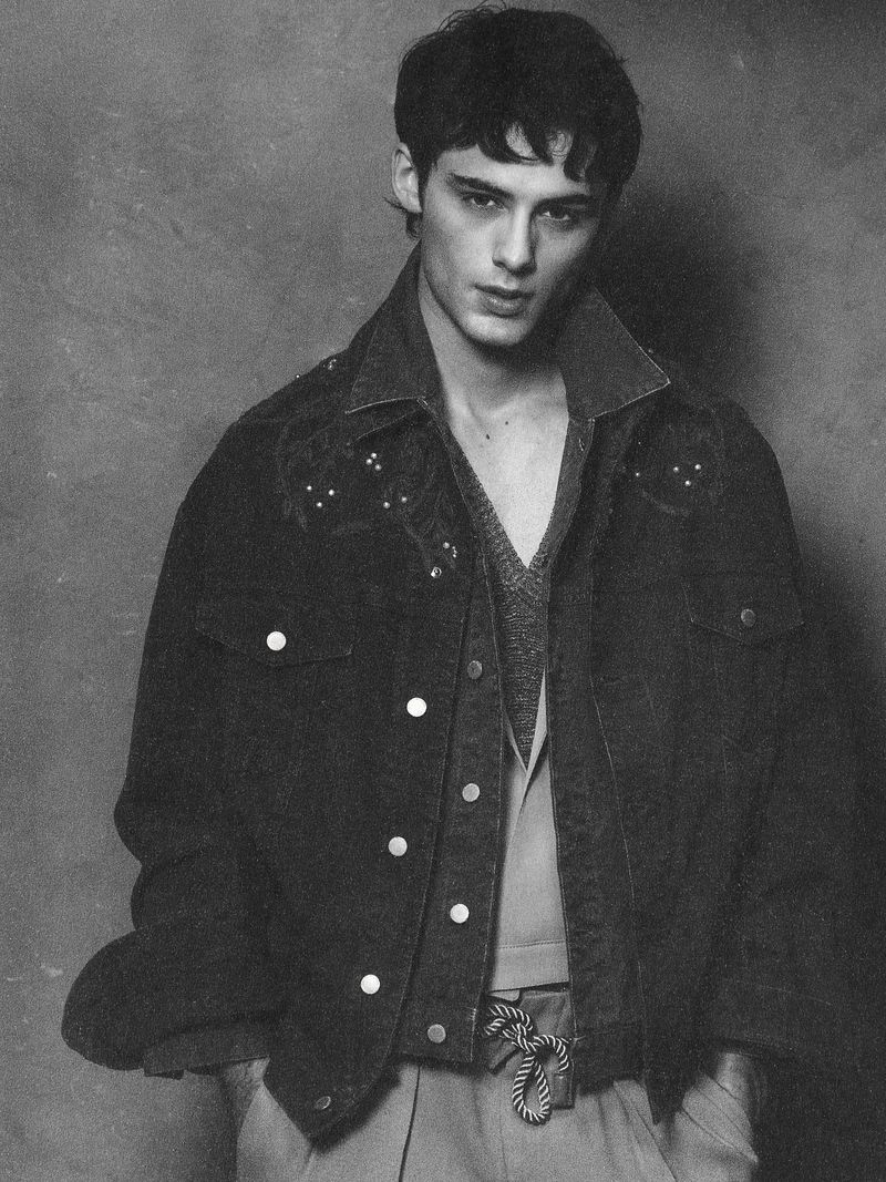 Black and white portrait of a young adult man wearing a denim jacket and vest, posing in front of a grey textured backdrop.