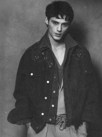 Black and white portrait of a young adult man wearing a denim jacket and vest, posing in front of a grey textured backdrop.