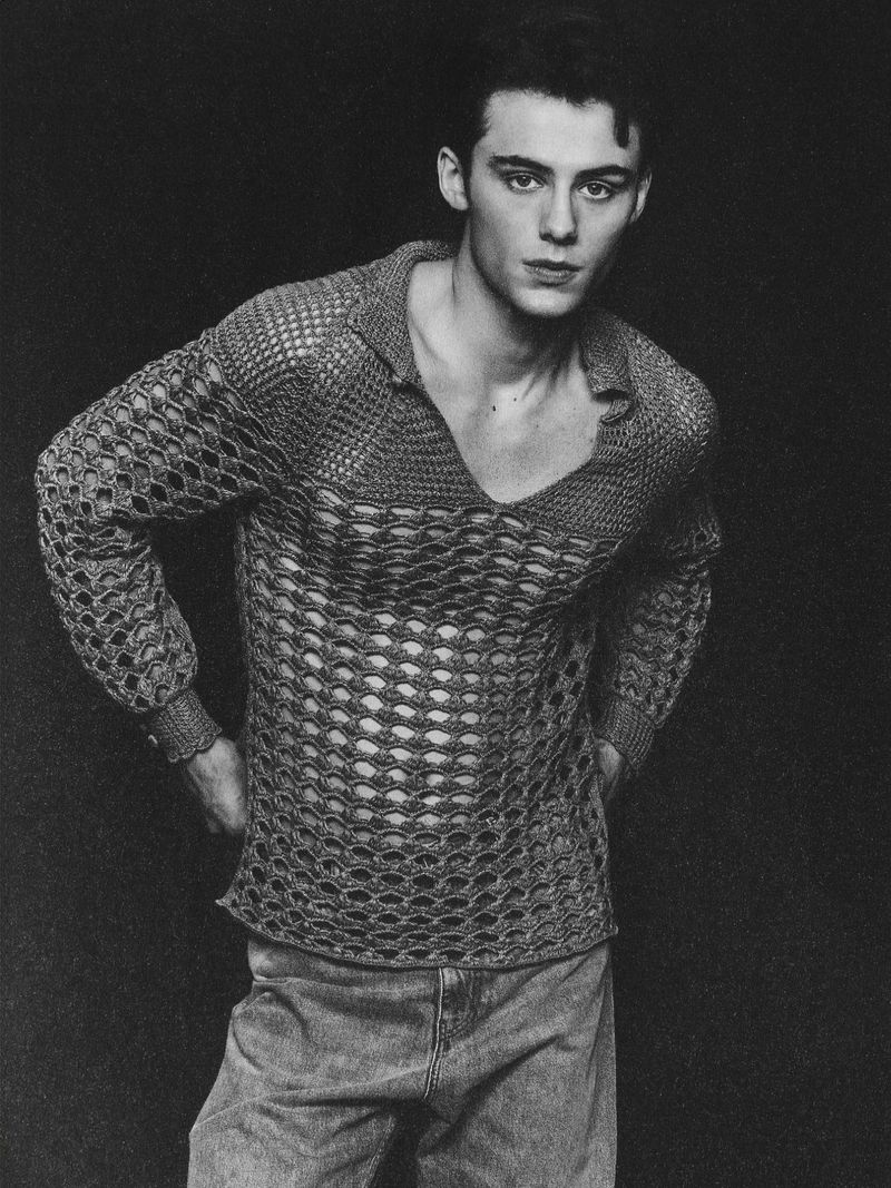 William Franklyn Miller wearing an open-weave crochet polo sweater and denim jeans against a black studio backdrop.