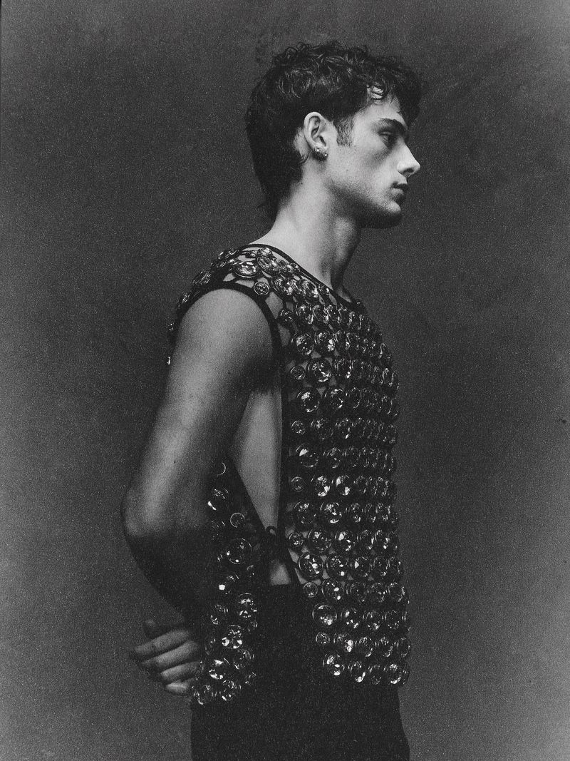 Black and white portrait of a young adult male in an embellished sleeveless shirt, looking off to the side in an indoor setting.