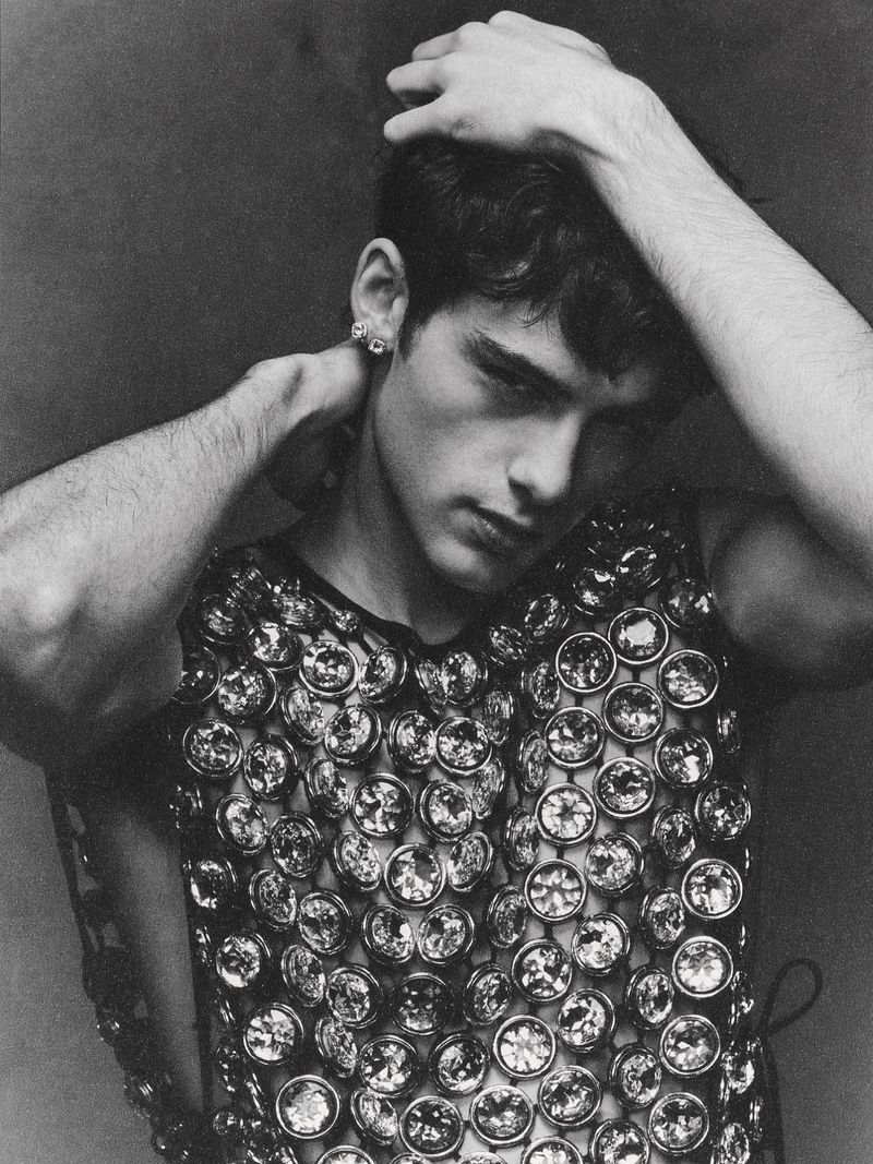 Black and white portrait of a young male model wearing a mesh top covered in round jewels, with his hands behind his head.