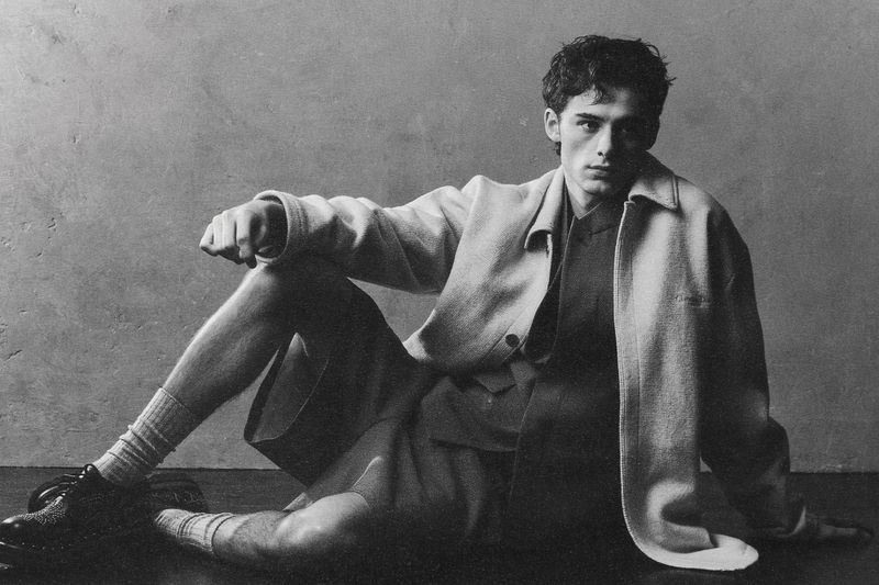 William Franklyn Miller seated on the floor wearing an oversized jacket, dark shorts, and dress shoes in a monochrome studio portrait.