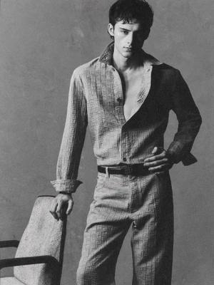 Monochrome portrait of young male model wearing patterned collared shirt and trousers standing beside a chair.