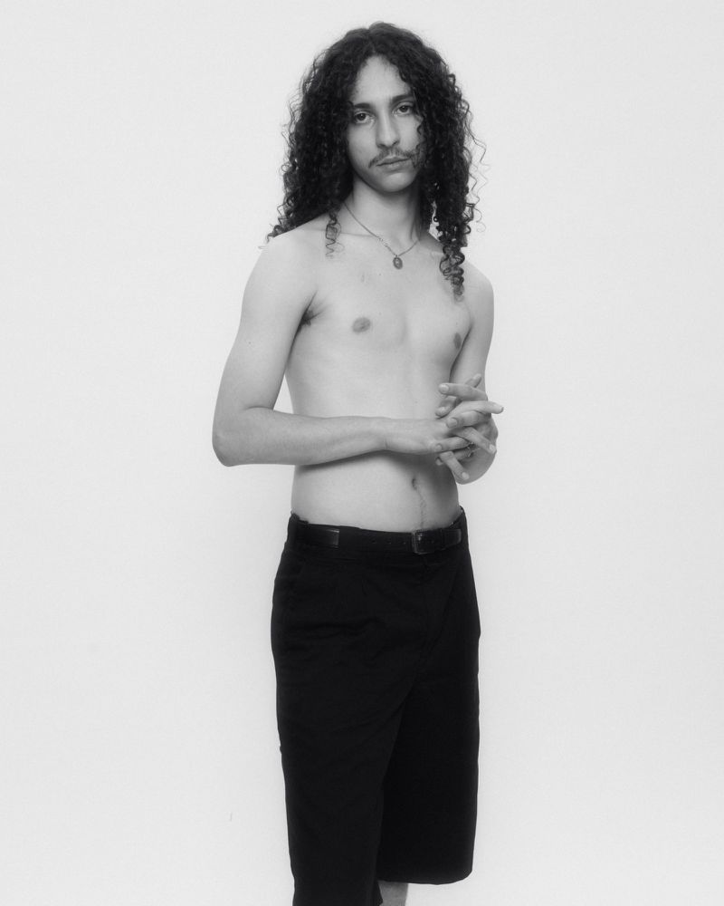 Portrait of a man with long curly hair and a mustache wearing dark trousers against a plain white studio backdrop.