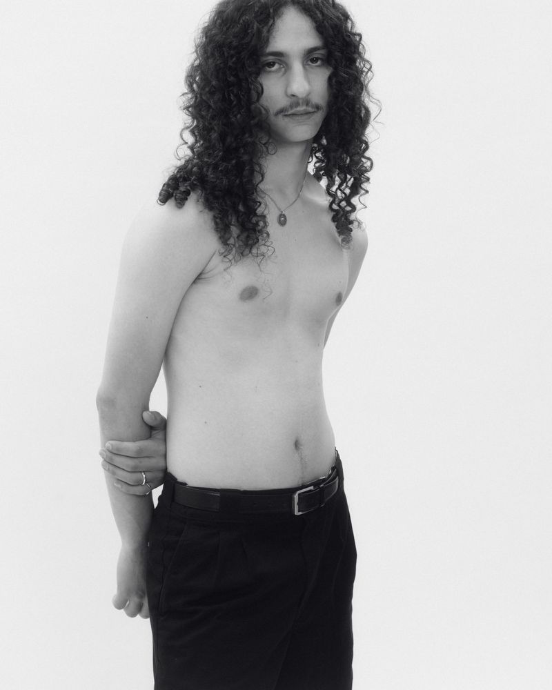 Black and white portrait of a shirtless adult male with curly hair and a mustache, wearing dark pants and a belt, posing indoors.
