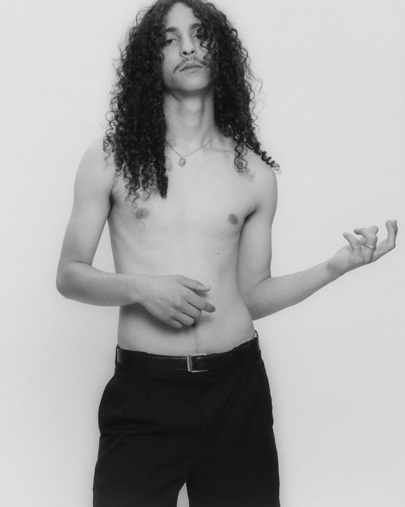 Portrait of a shirtless man with long curly hair and a mustache wearing black trousers against a light background.