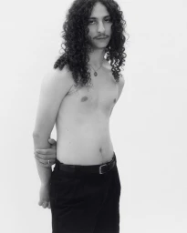 Portrait of a shirtless man with long curly hair and mustache wearing black trousers against a plain white background.