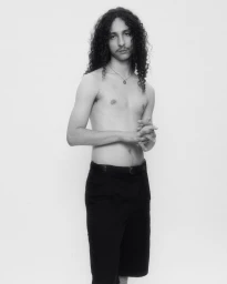 Portrait of a man with long curly hair and a mustache wearing dark trousers against a plain white studio backdrop.