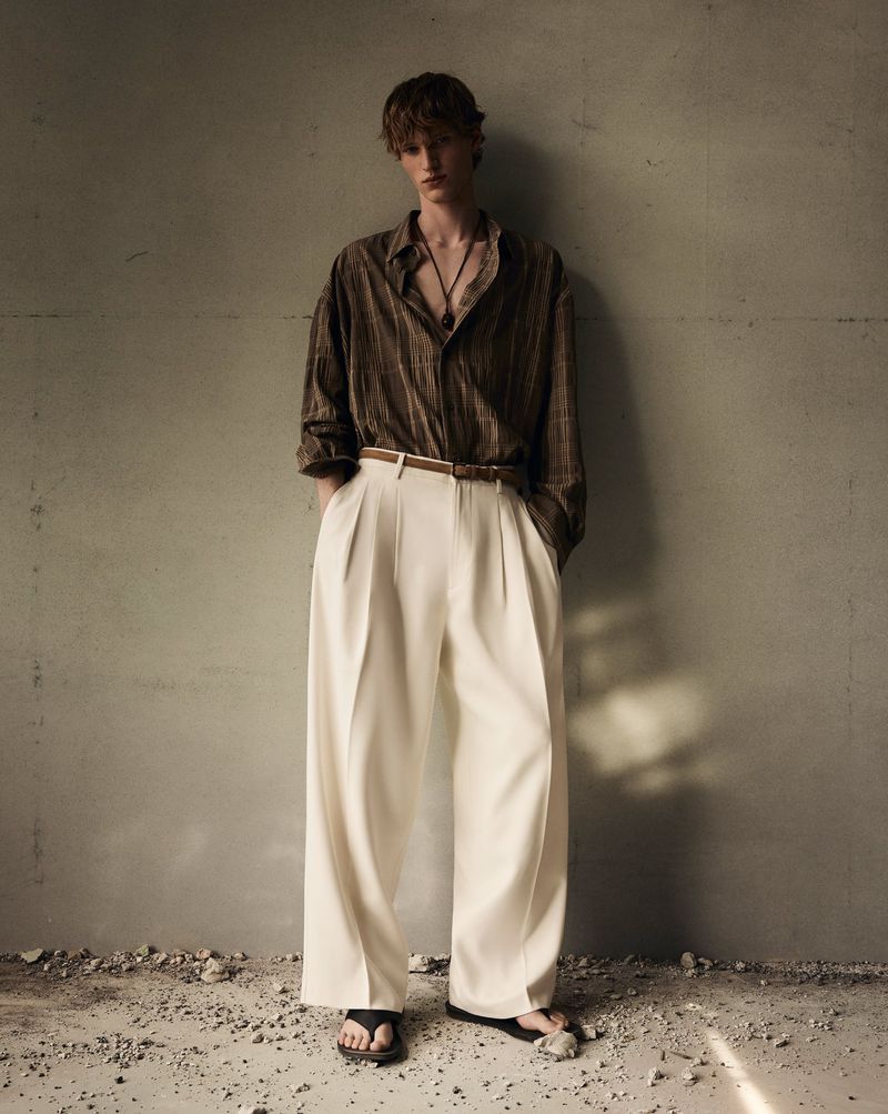 Full-length portrait of a male model wearing a striped brown shirt and wide white trousers leaning against a textured grey wall