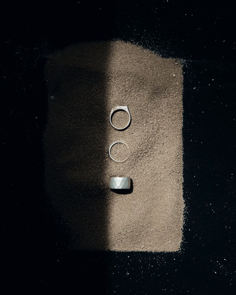 Three silver rings resting on a patch of brown sand bisected by a sharp vertical shadow against a dark background