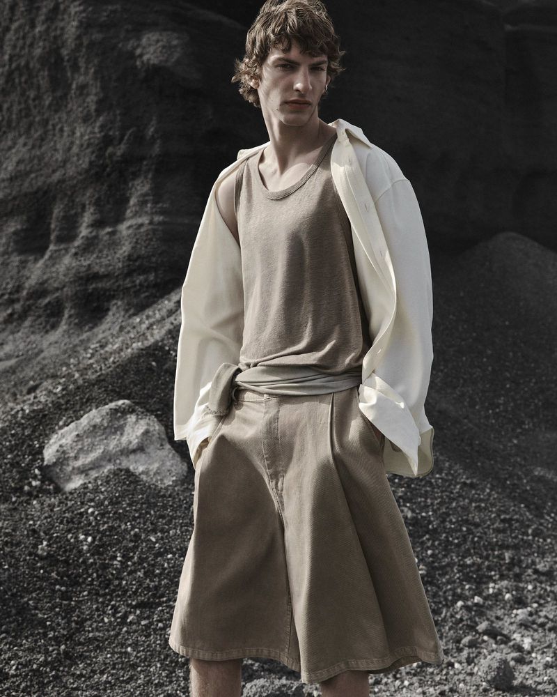Male model in a beige tank top and pleated shorts with an unbuttoned white shirt standing against dark rock formations.