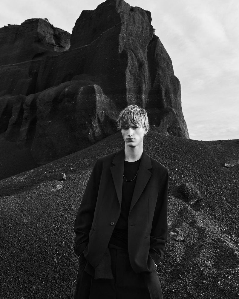 A young male model posing outdoors wearing a dark blazer in front of a massive dark rock formation and volcanic terrain.
