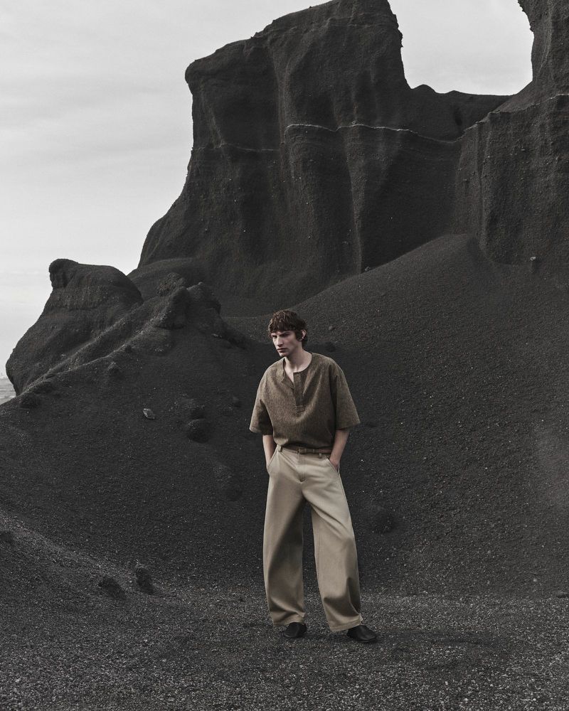 A male model wearing a linen shirt and trousers stands before towering dark rock formations.