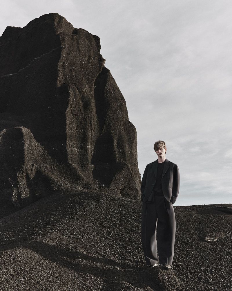 Model standing on a dark rocky terrain next to a towering volcanic rock formation under an overcast sky for Zara