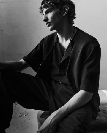 Black and white portrait of a male model wearing dark short-sleeved outerwear against a plain wall.