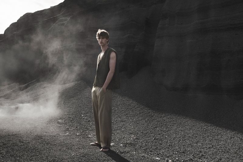 Model standing outdoors wearing an olive green sleeveless top and beige trousers in front of a dark rocky background.