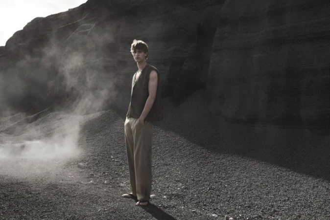 Model standing outdoors wearing an olive green sleeveless top and beige trousers in front of a dark rocky background.