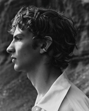 Profile portrait of a young man with curly dark hair wearing a white collared shirt and an ear cuff against a rocky background