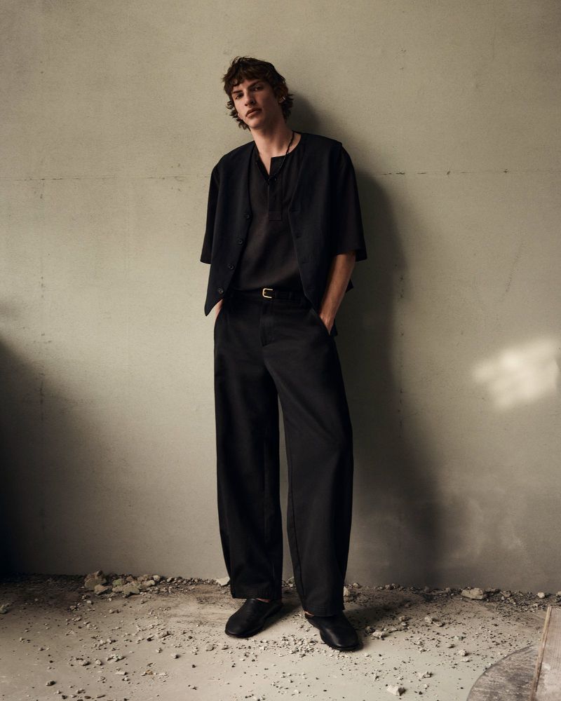 Full-length portrait of a male model leaning against a textured concrete wall wearing a dark shirt, trousers, and jacket.