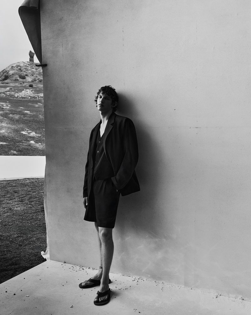 Monochrome fashion campaign image featuring a male model standing against a pale wall outdoors in a dark tailored suit and shorts.