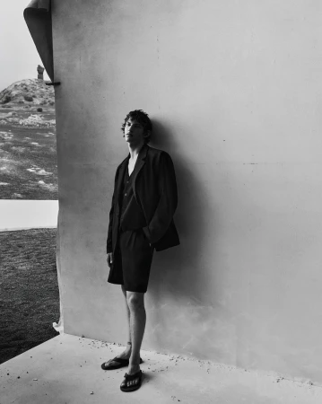 Monochrome fashion campaign image featuring a male model standing against a pale wall outdoors in a dark tailored suit and shorts.