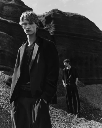 Two male models in dark attire posing outdoors against rugged rocky terrain in black and white photography.