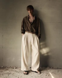 Full-length portrait of a male model wearing a striped brown shirt and wide white trousers leaning against a textured grey wall