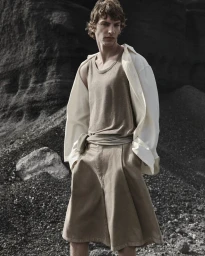 Male model in a beige tank top and pleated shorts with an unbuttoned white shirt standing against dark rock formations.