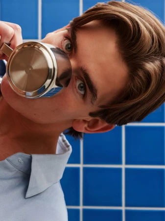 Close-up portrait of a young man holding a silver pitcher near his face against blue tiled background