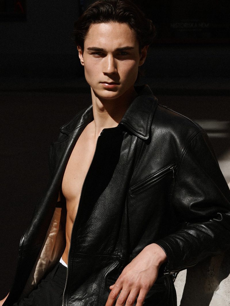 Young man with dark hair wearing an open black leather jacket outdoors with stark lighting casting shadows across his face