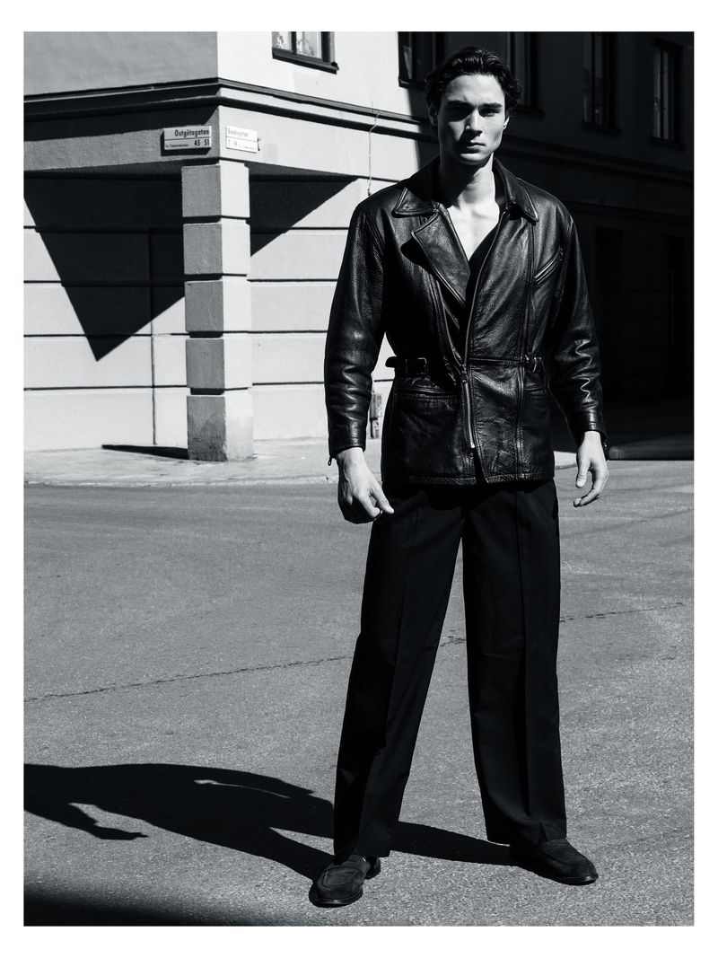Man wearing a dark leather jacket and trousers standing outdoors against an architectural background