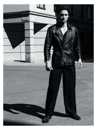 Man wearing a dark leather jacket and trousers standing outdoors against an architectural background
