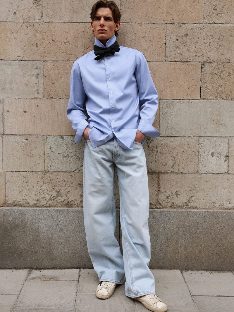 Man wearing a light blue button-up shirt, a black bow tie, and light blue denim jeans leaning against a stone wall.