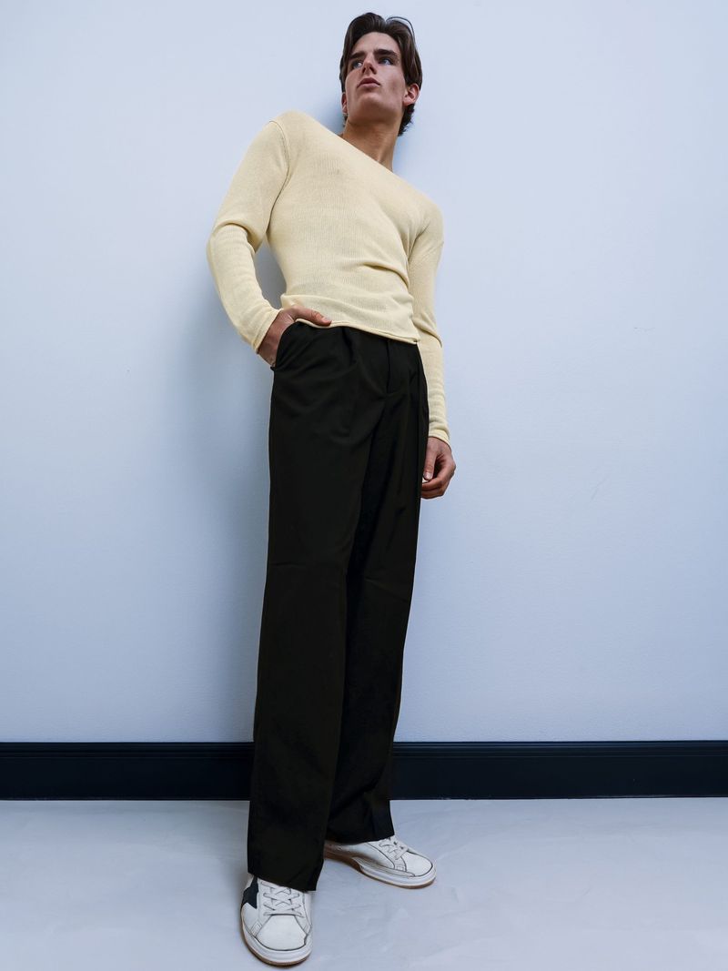 A young man in casual fashion poses in a photo studio with cream sweater, dark pants, and off-white sneakers, against a light blue wall.