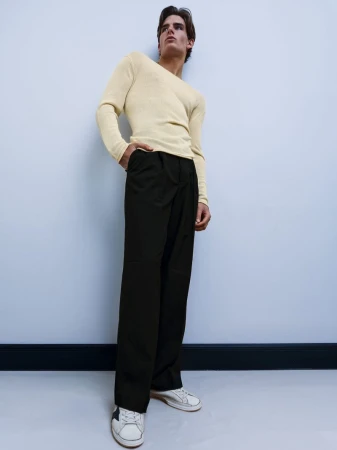 A young man in casual fashion poses in a photo studio with cream sweater, dark pants, and off-white sneakers, against a light blue wall.