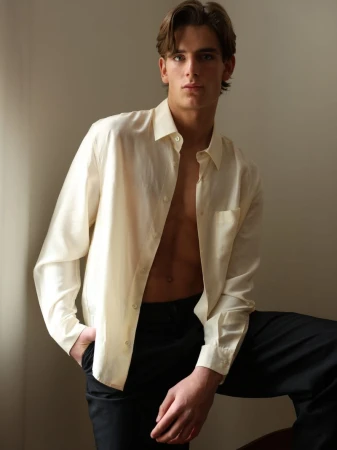 Young male model wearing an unbuttoned cream shirt and dark trousers against a neutral background