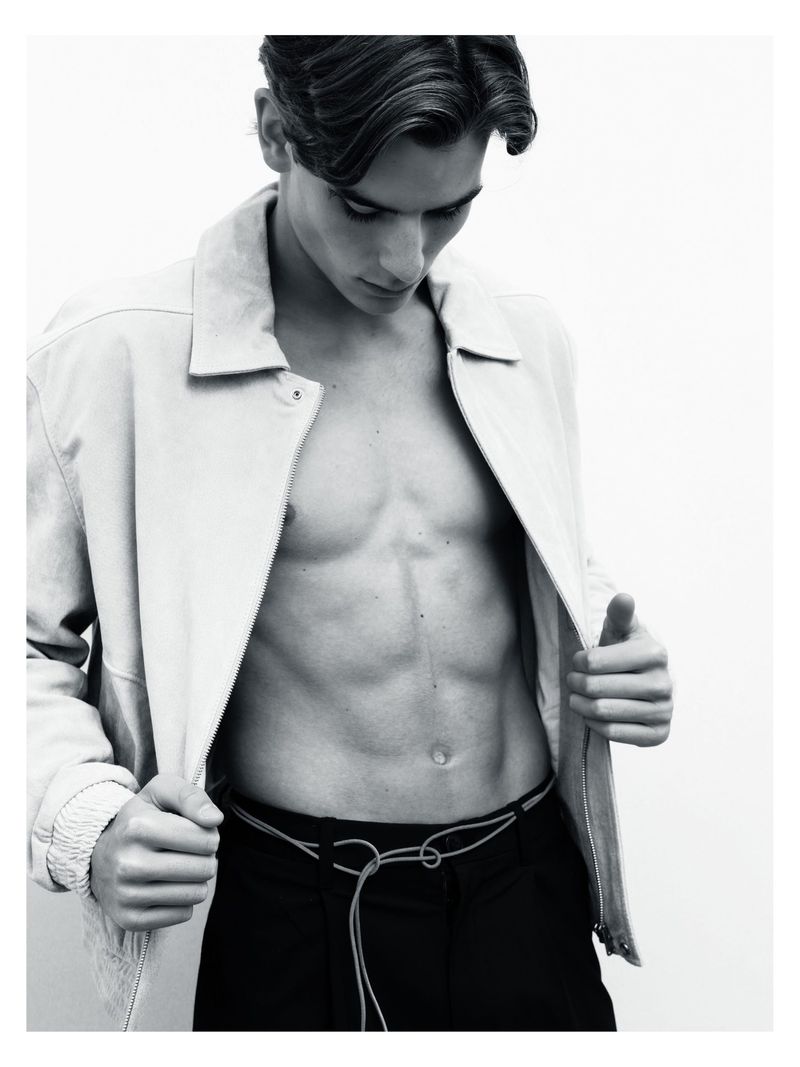 Shirtless male model wearing an open light jacket and dark trousers against a white seamless background