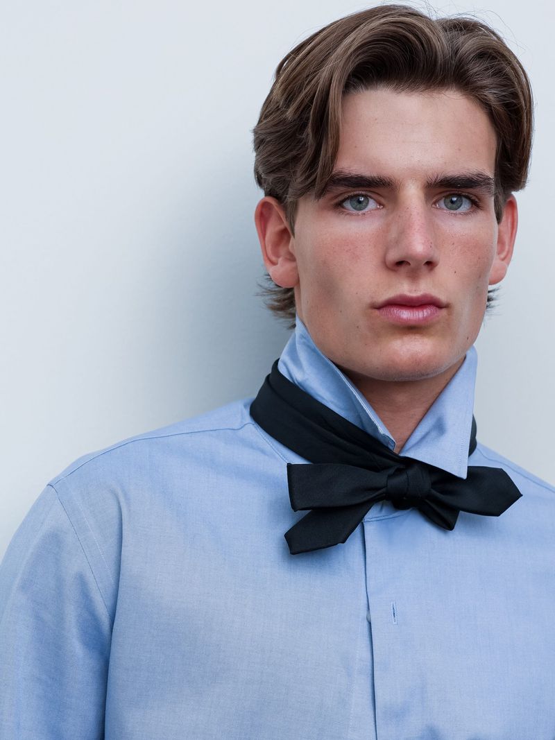 Close-up portrait of a young man wearing a light blue button-down shirt and a black bow tie against a white background.