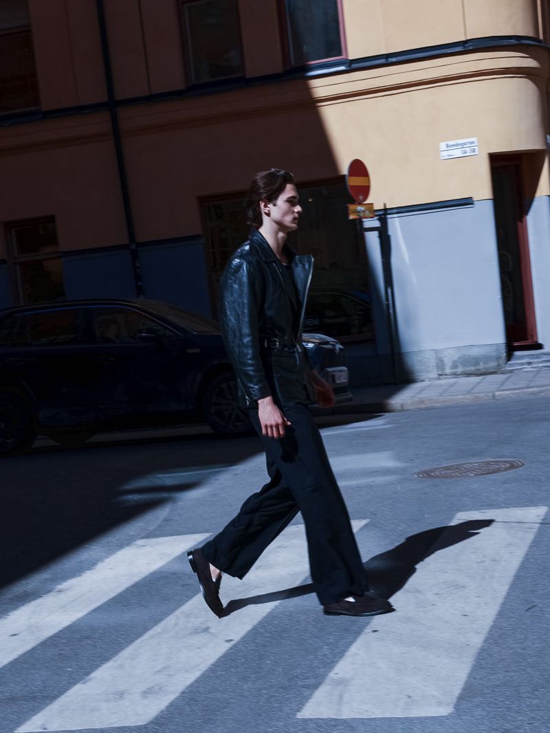 A man wearing a black leather jacket and dark trousers walking across a pedestrian crosswalk on a city street.