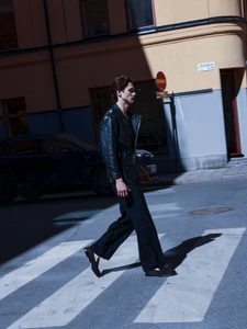 A man wearing a black leather jacket and dark trousers walking across a pedestrian crosswalk on a city street.