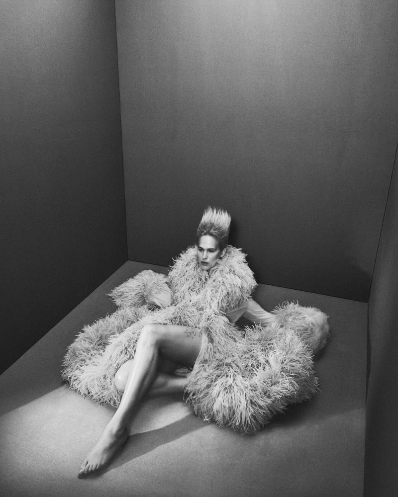Tommy Dorfman seated inside a dark grey corner studio wearing a feathered coat for Financial Times HTSI Magazine