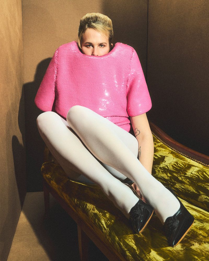 Tommy Dorfman wearing a structured pink top with legs tucked up on a velvet green set piece