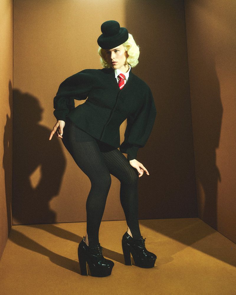 Tommy Dorfman posing in a dark green structured jacket, bowler hat, and platform shoes inside a brown set-dressed studio.