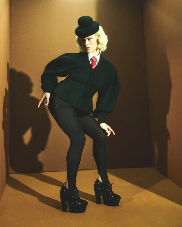 Tommy Dorfman posing in a dark green structured jacket, bowler hat, and platform shoes inside a brown set-dressed studio.