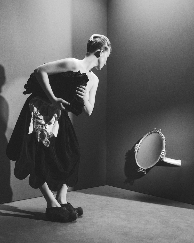 Monochrome portrait of a person in a dark gown looking toward an oval mirror held by hands emerging from a wall.