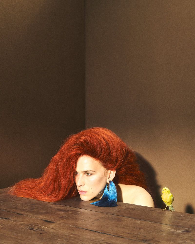 Tommy Dorfman with long red hair and a blue tassel earring resting on a wooden surface beside a small yellow bird