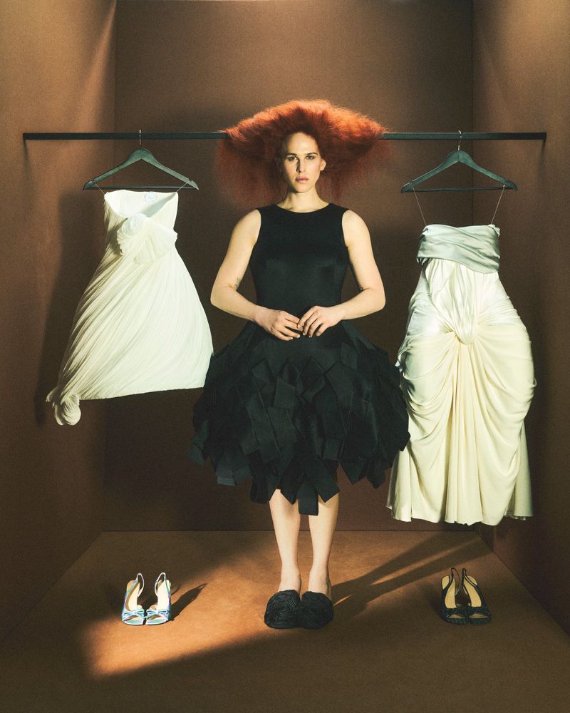 Tommy Dorfman with voluminous red hair wearing a black dress flanked by two dresses on hangers for Financial Times HTSI Magazine