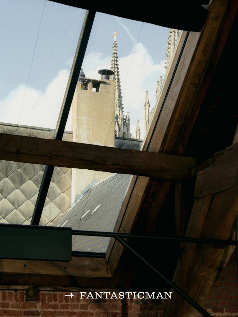 Architectural view through a skylight showing weathered brick, exposed timber beams, and a church spire against a blue sky.