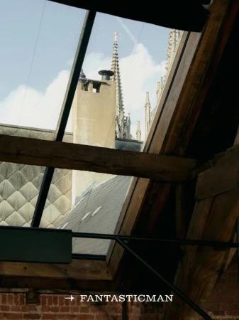 Architectural view through a skylight showing weathered brick, exposed timber beams, and a church spire against a blue sky.